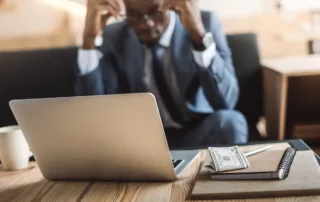 A man in a blue suit is sitting on a sofa in the blurred background, looking down with his hands near his head, conveying stress or distress. In the foreground, on a wooden table, a laptop, a notebook with a pen, and a stack of US dollar bills are visible. The image suggests financial problems, debt, or difficult decisions, possibly related to divorce or bankruptcy.