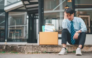 Worried man in a blue shirt and tie sits on a step outside an office, with a box of belongings and a resignation letter by his side, symbolizing job loss and financial uncertainty.