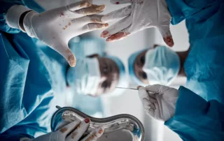 Surgeons in blue scrubs and masks operate from the patient’s perspective, with stained gloved hands visible, conveying intensity and surgical risk.