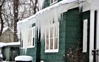 Icicles hanging from the roof of a green house in snowy West Virginia, with bare trees—illustrating winter hazards and property owner responsibilities for safety.